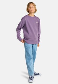 Sweat-shirt violet à encolure ronde avec logo brodé, porté avec un jean denim bleu clair et des baskets bleu marine.
