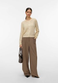 Cardigan en maille beige clair avec un motif de cœur, présentant un col rond et une fermeture à boutons, associé à un pantalon marron à jambes larges.