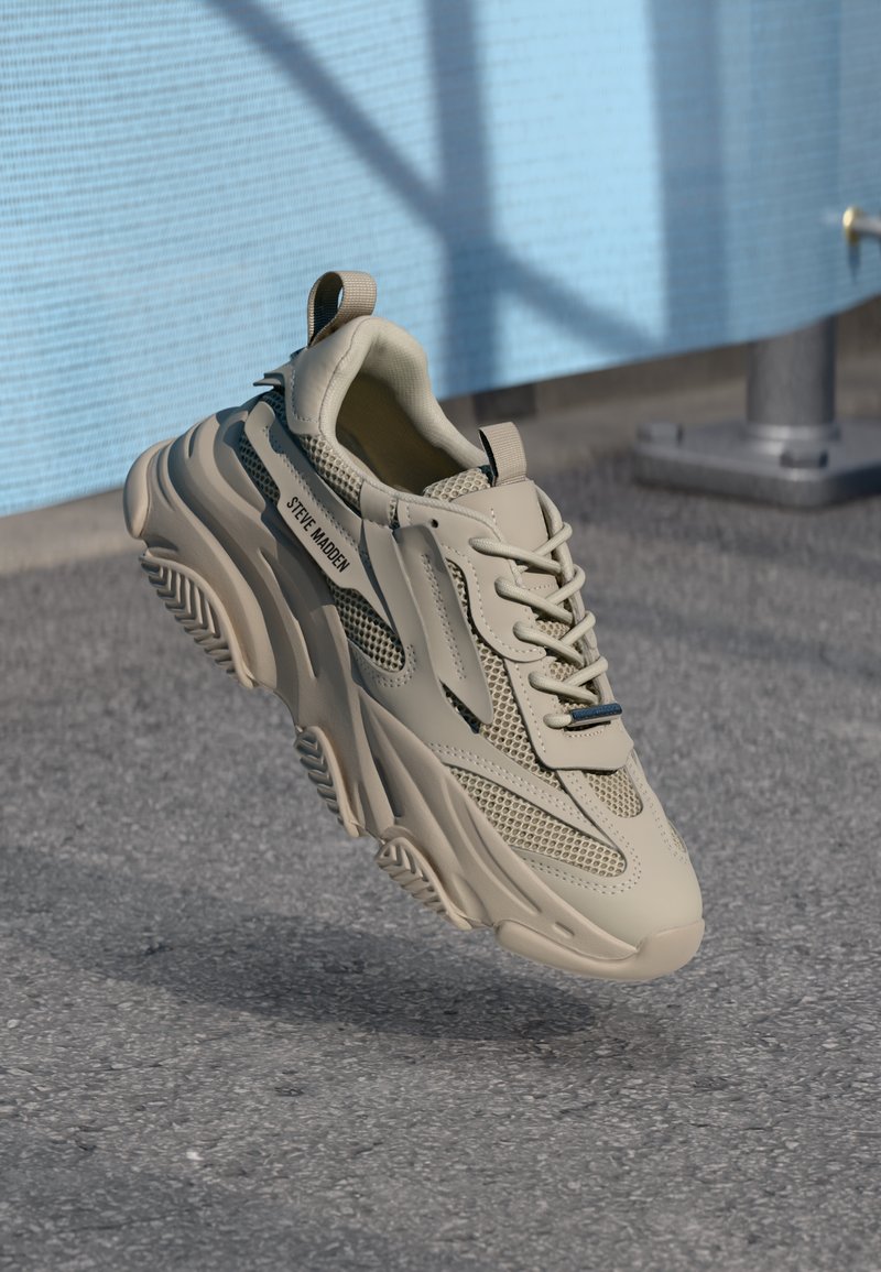 Baskets beiges Steve Madden avec une semelle épaisse et des détails en mesh, flottant au-dessus d’un sol texturé près d’un mur bleu et d’un poteau en métal.