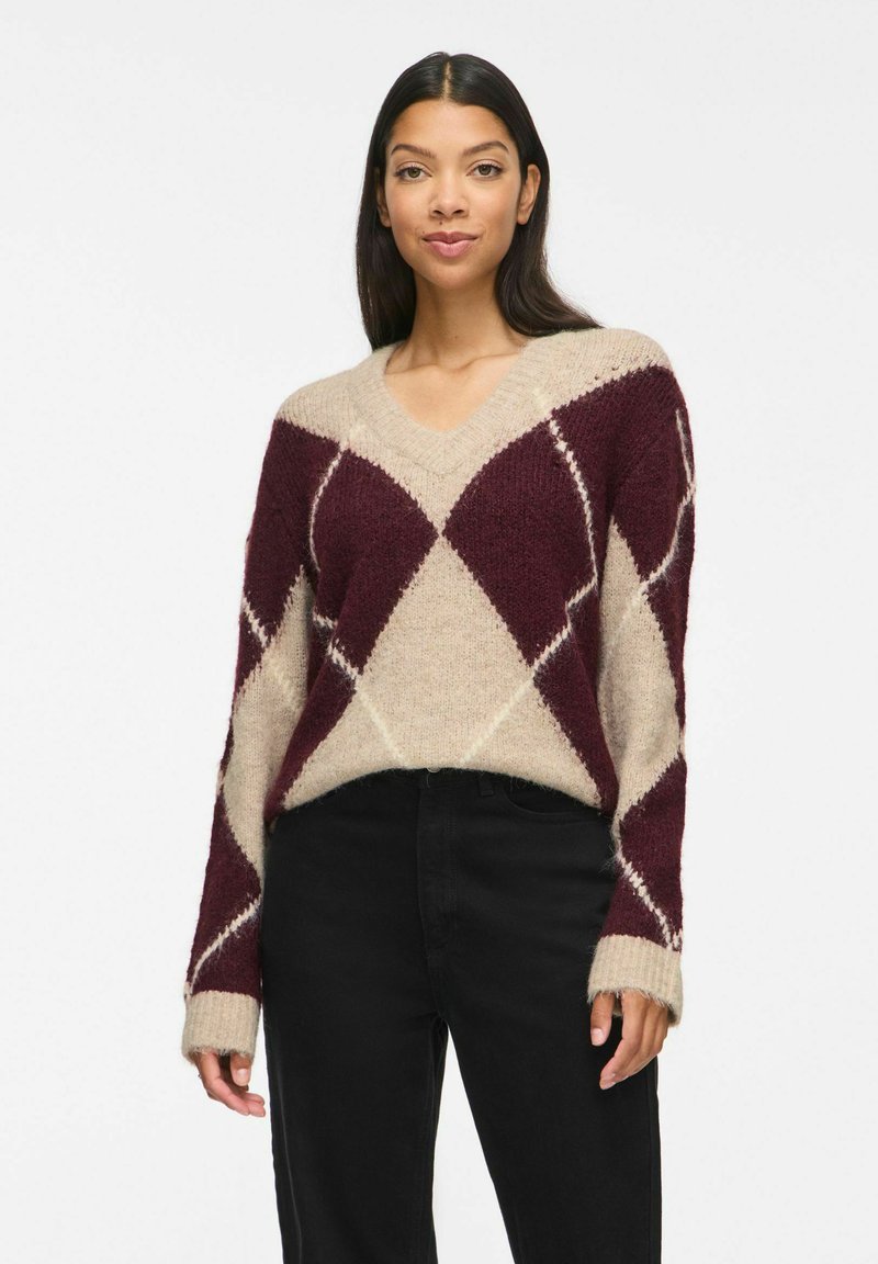 Femme aux longs cheveux foncés portant un pull beige et bordeaux à col en V à motif losanges et un pantalon noir, debout devant un fond blanc.