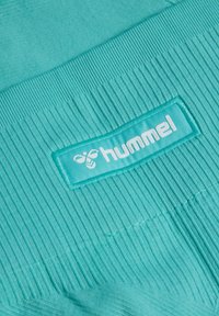 Turfkleurige sportkledingstof met ribbelstructuur, voorzien van een lichtblauwe rechthoekige patch met het "hummel"-logo in witte letters.