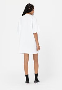 Biała, oversize sukienka t-shirtowa z krótkimi rękawami, gładka bawełniana tkanina, boczne rozcięcia, noszona z czarnymi skarpetkami i czarnymi trampkami.