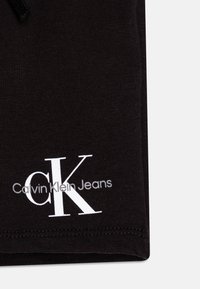 Čierna bavlnená látka so snehobielym logom "Calvin Klein Jeans" a štýlizovaným "CK" v jednoduchom písmovom písme, s hladkou textúrou.