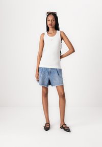 Hvit ermeløs tank topp med en liten logo, kombinert med lyseblå denimshorts med en råkant og subtil tekstmønster. Svart flate sko.