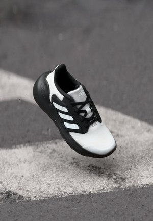 Zapatilla deportiva en blanco y negro con parte superior de malla, tres franjas blancas a un lado y un cuello acolchado. Suela texturizada para tracción.