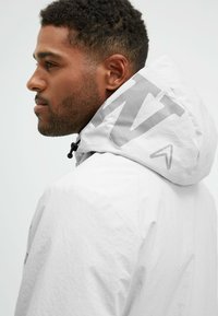 Chaqueta ligera blanca con capucha gris contrastante que presenta un gran logo. La tela parece suave con un ligero brillo. Cuerdas ajustables en la capucha.