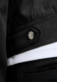 Czarna zamszowa kurtka z srebrnym zatrzaskiem, noszona na białej koszuli i czarnych spodniach w prążki. Prezentuje fakturowaną tkaninę i detale przeszyć.