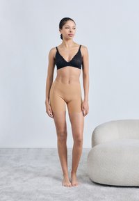 Μαύρο λευκό δαντελένιο σουτιέν με μαλακό ντεκολτέ σε συνδυασμό με καφέ απαλά, εφαρμοστά σορτς. Το μοντέλο ποζάρει μπροστά σε ανοιχτόχρωμο φόντο πάνω σε μια υφασμάτινη μοκέτα.