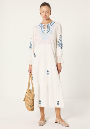 Γυναίκα που φοράει μακρύ λευκό φόρεμα με μπλε κέντημα, κρατάει υφαντή ψάθινη τσάντα και φοράει μπεζ υφασμένα φλατ παπούτσια, στέκεται σε μονόχρωμο φόντο.