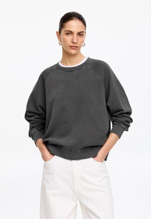 Kvinde med mørkt hår iført ørepiercinge og en løs mørkegrå sweatshirt samt hvide bukser med hænderne i lommerne mod en hvid baggrund.