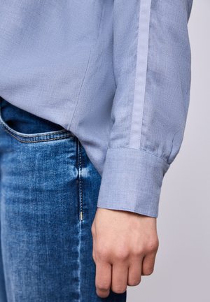 Detalje af kant og ærme på en lyseblå skjorte med lange ærmer over blå denimjeans, med hånden afslappet ved siden af kroppen.