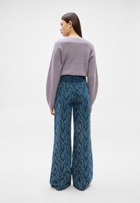 Γυναίκα με μακριά σκούρα μαλλιά φοράει ένα πλεκτό πουλόβερ λεβάντας και πλατύφρακτα μπλε τζιν με σχέδιο, στέκεται γυρισμένη πλάτη σε λευκό φόντο.