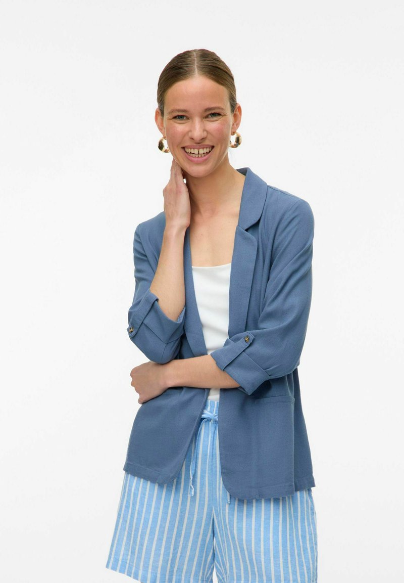 Šviesiai mėlynas blazeris su sukniukėmis, vienu mygtuku užsegimas ir tekstūruota medžiaga, derinamas su mėlynais ir baltais dryžuotais šortais.