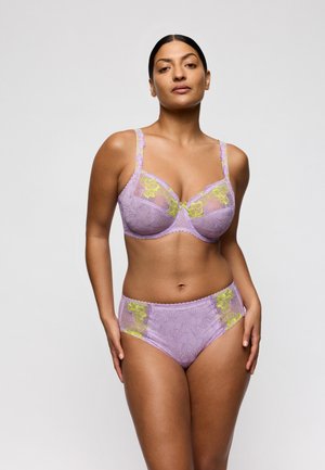 Vrouw draagt een lavendel kleurige kanten beha en bijpassende hooggetailleerde slip met gele bloemrijke borduursels, staand tegen een effen lichte achtergrond.