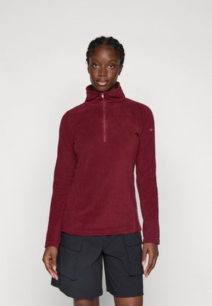 Kvinne iført en marsjvinsfarget fleece med kvart glidelås og svarte cargo-shorts, stående mot en lys ensfarget bakgrunn.