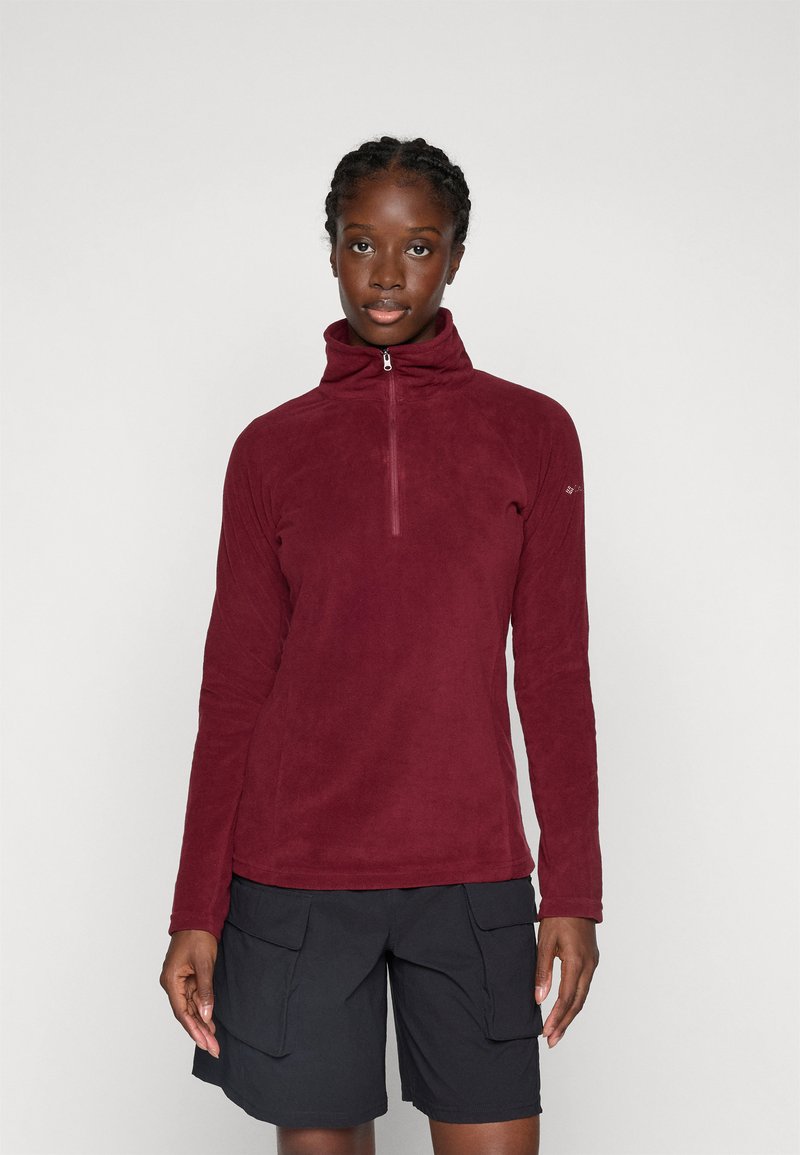 Vrouw in een maroon fleece met kwart rits en zwarte cargo shorts, staande tegen een effen lichte achtergrond.
