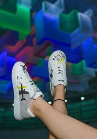 White sneakers with black airplane graphics and "NEW YORK" text, featuring a striped design and rounded toe, set against a colorful background.