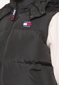 Schwarze Steppweste mit Reißverschluss, gepolsterter Textur und Kapuze. Verfügt über ein kleines Tommy Jeans Logo-Patch auf der Brust.