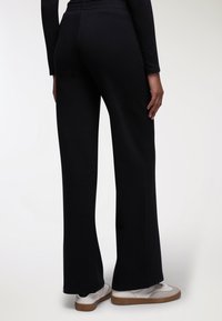 Pantaloni di felpa neri a maglia con un design a gamba svasata, dotati di una cintura testurizzata e abbinati a sneaker bianche e beige.