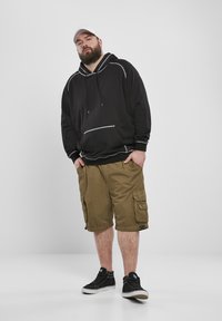 Felpa nera con cappuccio e dettagli bianchi, pantaloni cargo khaki con tasche laterali e sneakers nere. Cappellino grigio indossato al contrario.