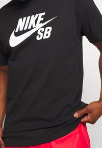 Osoba v černém tričku Nike SB a červených šortkách, s rukou držící okraj trička.
