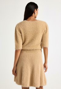 Beige fluffig stickad tröja med korta ärmar, i kombination med en matchande utsvängd kjol. Mjuk textur och en svag lyster synlig på tyget.