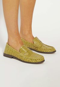 Green faux leather loafers with a textured, croc-like pattern, slip-on design, and a flat brown sole.