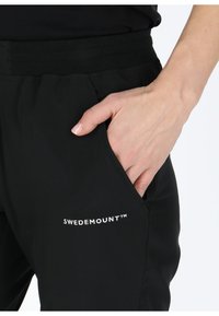 Pantaloni sportivi neri con cintura elastica, dotati di una tasca laterale e logo "SWEDEMOUNT™" ricamato sulla gamba inferiore.