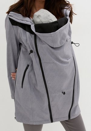 Femme portant un manteau de portage gris avec un bébé capuchonné dans la poche avant, montrant les poches zippées et les cordons de serrage réglables.