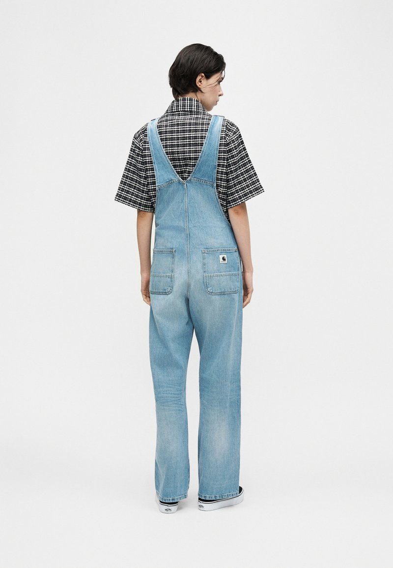 Personne de dos portant une salopette en denim bleu clair et une chemise à manches courtes à carreaux noir et blanc, debout devant un fond blanc uni.