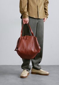 Brown leather bag with a round shape, soft texture, and dual handles. Paired with gray trousers and woven light shoes.
