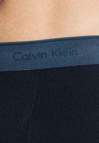 Schwarze Baumwollboxershorts mit einem marineblauen elastischen Bund, der das geprägte Logo "Calvin Klein" trägt. Nahtdetails sind auf dem Stoff sichtbar.