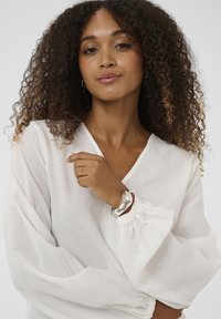 Femme aux cheveux bouclés portant un chemisier blanc et un large bracelet argenté, posant avec la main près de sa poitrine sur un fond uni.