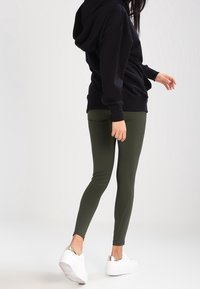 Felpa con cappuccio nera e leggings verde oliva abbinati a sneaker bianche con dettagli metallici. Tessuto morbido e design aderente.