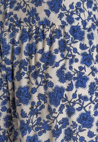 Tissu floral avec des motifs bleus sur un fond beige, présentant des motifs et textures élaborés. Le matériau semble doux avec un léger éclat.