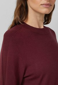 Frau trägt einen dunkelroten Strickpullover mit gerippten Schultern, der den Hals und das untere Gesicht vor einem schlichten grauen Hintergrund zeigt.