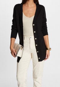 Cardigan noir en maille, avec un col en V, des manches longues et des boutons métalliques. Associé à un pantalon de couleur crème et un sac à main.