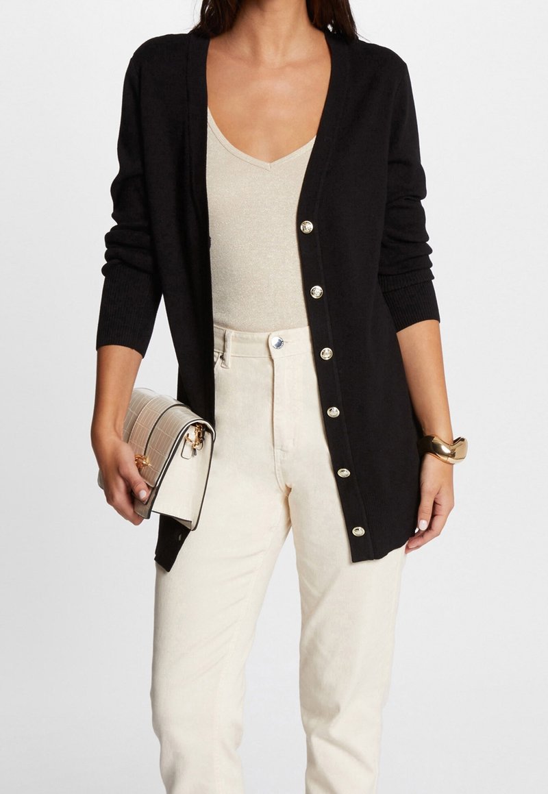 Cardigan noir en maille, avec un col en V, des manches longues et des boutons métalliques. Associé à un pantalon de couleur crème et un sac à main.