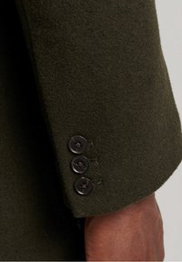 Manche de veste en laine vert olive avec trois boutons foncés, tissu texturé et coutures décoratives à la jonction entre le poignet et le bras.