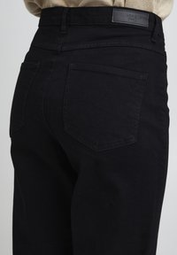 Jean en denim noir à taille haute, avec deux poches arrière et une étiquette logo noire sur la ceinture. Texture lisse et coupe ajustée.