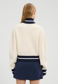 Kremowy sweter w prążki z granatowym kołnierzykiem i mankietami, z białymi paskami akcentującymi. Nosi się go na granatowej spódnicy, eksponując dopasowany krój.