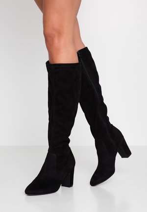 Bottes noires en suède montantes jusqu'aux genoux avec talons carrés portées sur des jambes croisées sur un fond clair uni.
