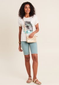 Weißes T-Shirt mit einem Blatt-Graphic, helltürkisfarbene Shorts und eine goldene Umhängetasche. Das Model trägt beigefarbene Sandalen und hebt ein lässiges Sommeroutfit hervor.