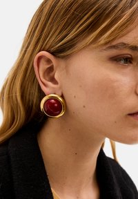 Náušnice ve zlatém odstínu s velkým kulatým červeným kamenem, zasazeným v hladkém, kulatém rámečku, vytvářející odvážný a lesklý vzhled.