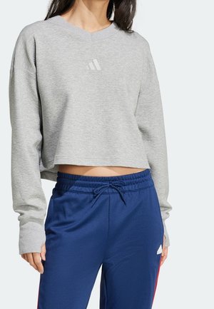 Femme portant un top court côtelé gris clair à manches longues et un pantalon bleu à cordon avec un petit logo sur la poitrine et la jambe.