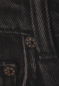 Close-up de tecido de ganga preta com um botão metálico preto gravado com a palavra "DAYWEEK" em letras cor de cobre.