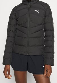 Černá bundy s prošíváním a zipem, s chevronovým vzorem a bílým logem Puma na levé hrudi, v kombinaci s tmavými šortkami.