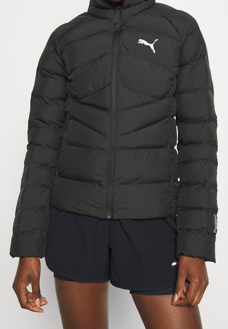 Černá bundy s prošíváním a zipem, s chevronovým vzorem a bílým logem Puma na levé hrudi, v kombinaci s tmavými šortkami.