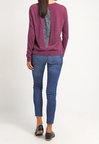 Bordowy sweter z dużym wykrojem na plecach, odsłaniającym wzorzystą tkaninę pod spodem. W zestawieniu z dopasowanymi niebieskimi dżinsami i czarnymi butami.