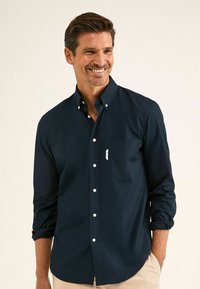 Camicia blu navy con colletto strutturato, maniche lunghe arrotolate ai polsi, tasca sul petto e bottoni bianchi. Tessuto dalla trama liscia.